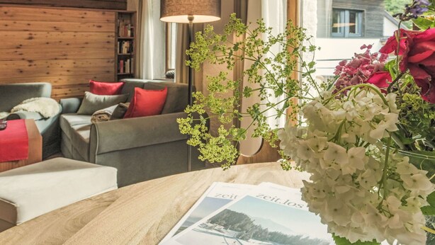 A cozy living room with a wooden ceiling, a sofa with red cushions, a table with a magazine, and a vase with flowers.