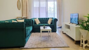 Un salon moderne avec un canapé d’angle vert, une table basse blanche et un téléviseur posé sur un meuble blanc.
