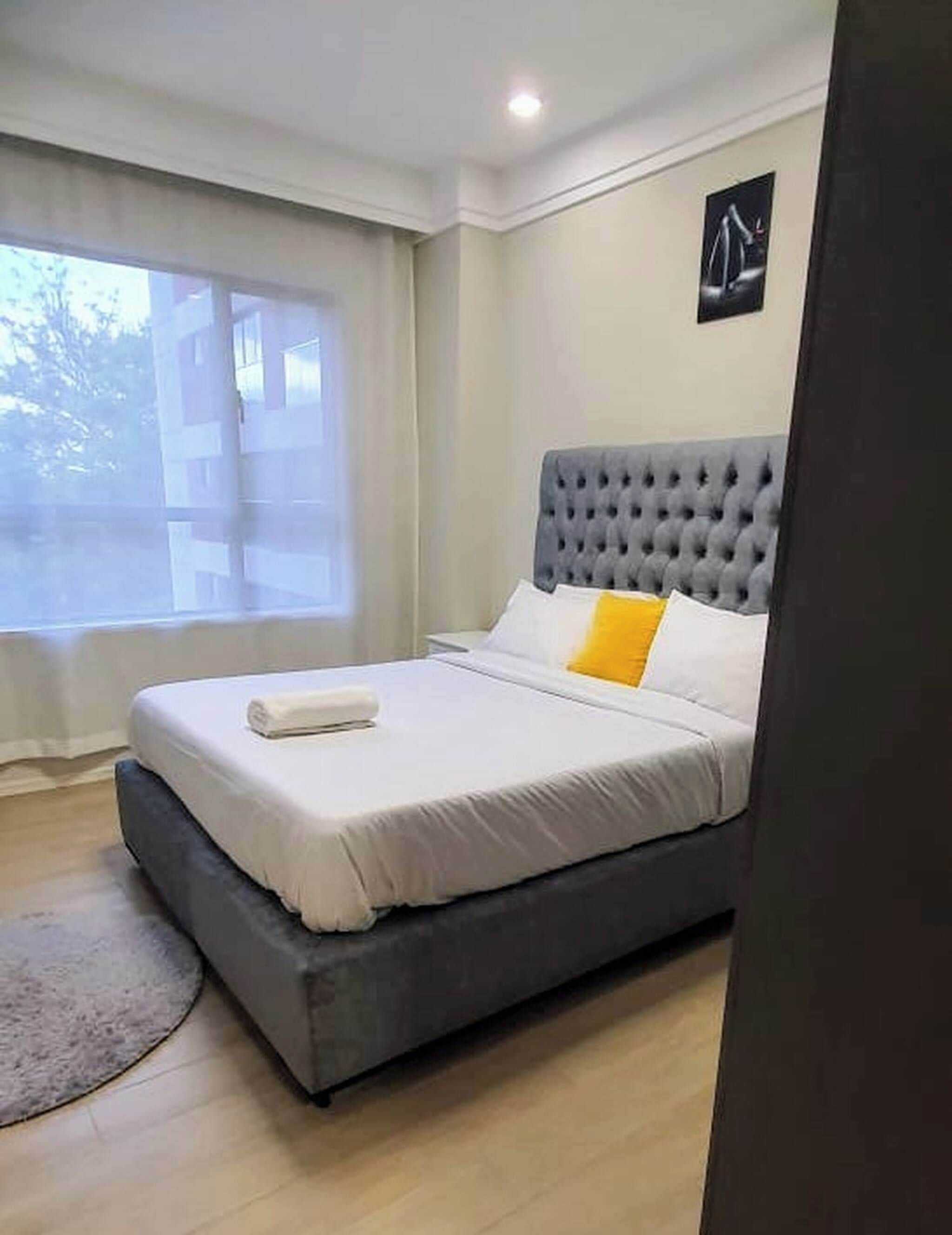 Quarto com cabeceira estofada cinza, roupa de cama branca, almofada amarela e janela com cortinas.