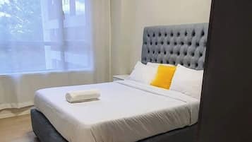 Quarto com cabeceira estofada cinza, roupa de cama branca, almofada amarela e janela com cortinas.