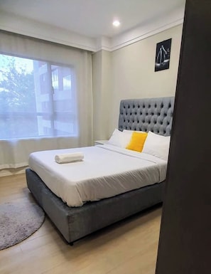 Een slaapkamer met een grijs gestoffeerd hoofdeinde, wit beddengoed, een geel kussen en een raam met gordijnen.