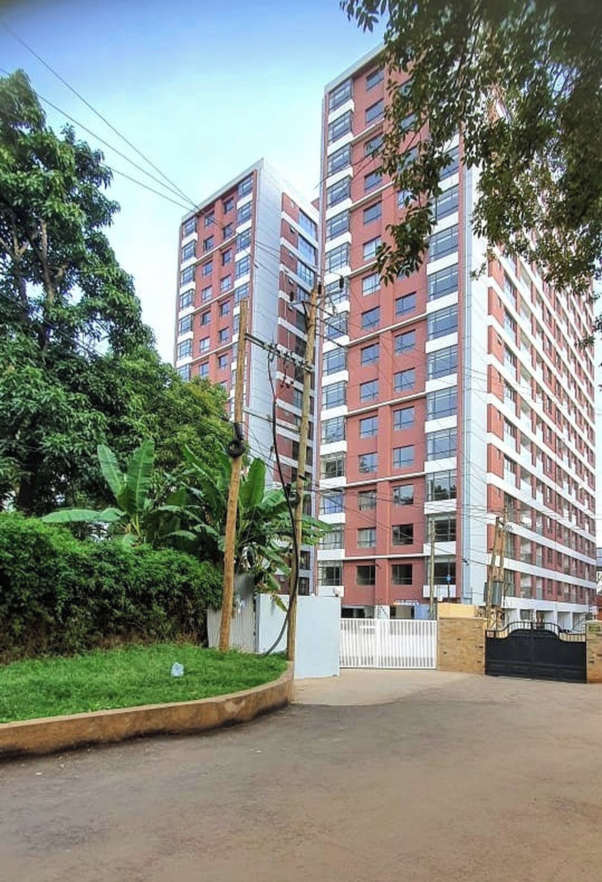 Um edifício residencial alto, com vários andares, rodeado por vegetação e um portão.