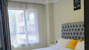Un lit bien fait, avec une tête de lit grise, une literie blanche et un seul oreiller jaune.