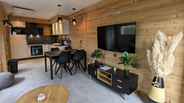 A modern kitchen with a built-in oven and microwave, a dining area with a wooden table and chairs, and a living area with a TV and decorative plants.
