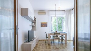 Una sala moderna con televisor, asientos, una mesa de comedor y un gran ventanal con cortinas.