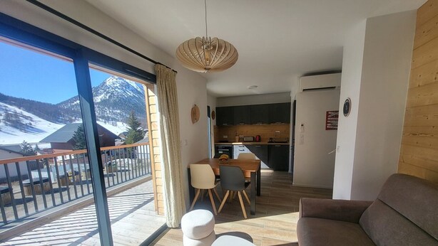 Un salon moderne avec un canapé, une table à manger et des chaises, une cuisine en arrière-plan et un balcon offrant une vue sur les montagnes.