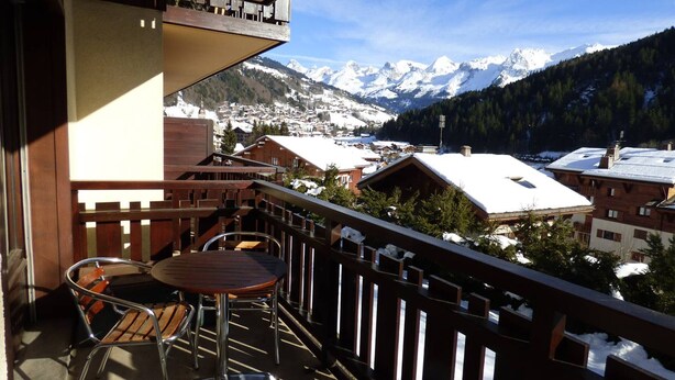 Ein Balkon mit Tisch und Stühlen, mit Blick auf eine verschneite Berglandschaft und Gebäude.
