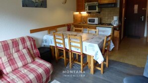 A cozy living room with a plaid sofa and a dining table set for four. A kitchen with wooden cabinets and a microwave is visible in the background.