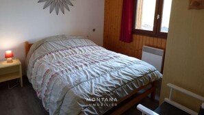 A bedroom with a bed, a nightstand with a lamp, a window with a curtain, and a wall-mounted decorative item.