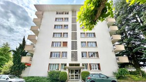 A multi-story building with a central entrance, balconies on each floor, and a parking area in front.