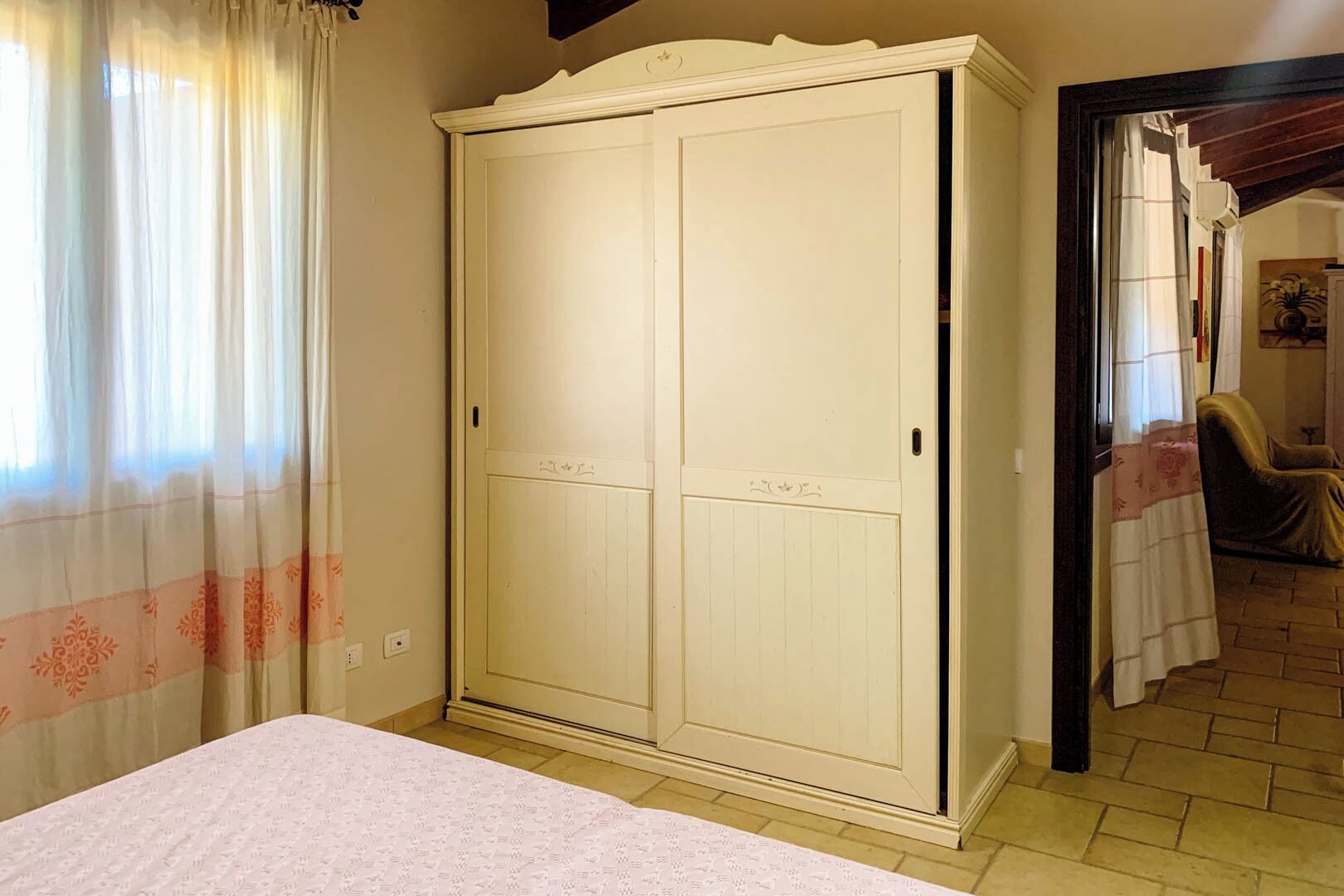 Un dormitorio con un armario grande, una cama con colcha estampada y una ventana con cortinas translúcidas.