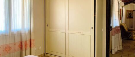 A bedroom with a large wardrobe, a bed with a patterned bedspread, and a window with sheer curtains.