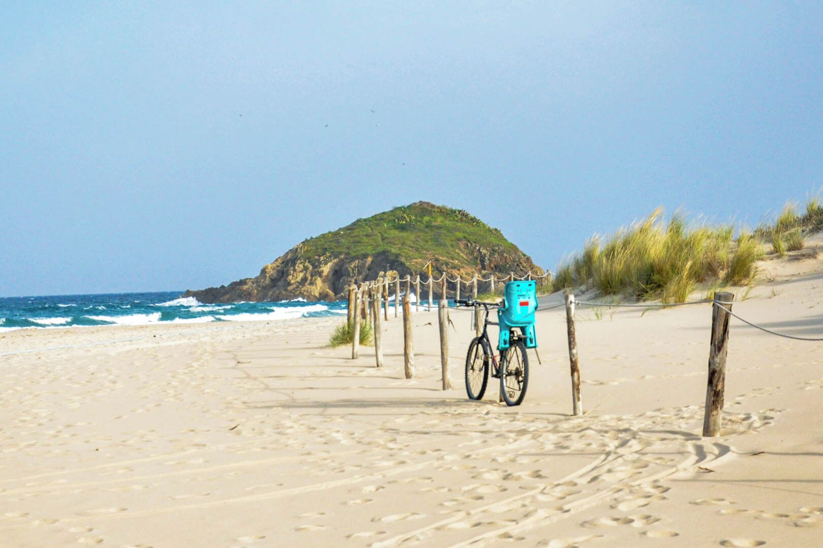 Una playa con una bicicleta y una cesta azul, un camino de arena y una colina rocosa al fondo.