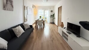 A modern living room with a black sofa, a dining table with chairs, a TV on a stand, and a large window with curtains. - Juan-les-Pins, Antibes - 3-bedroom apartment near beaches and town center (Antibes)