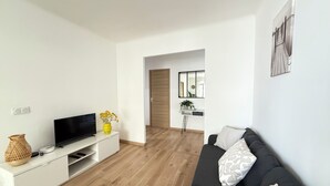A modern living room with a flat-screen TV on a white console, a black sofa with patterned cushions, and a wooden floor. - Juan-les-Pins, Antibes - 3-bedroom apartment near beaches and town center (Antibes)