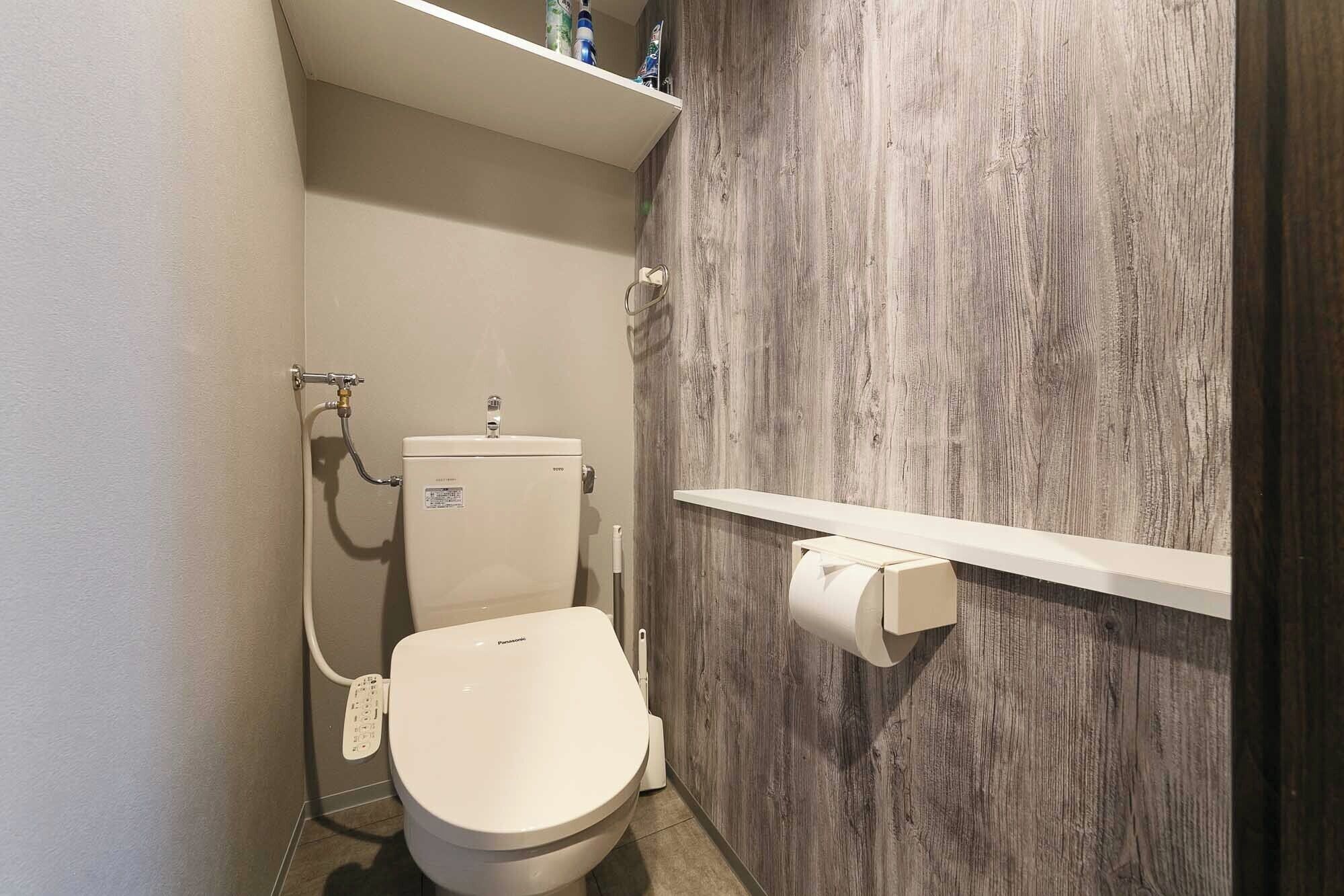 A compact bathroom with a white toilet, a wooden wall, and a shelf above the toilet holding toiletries.