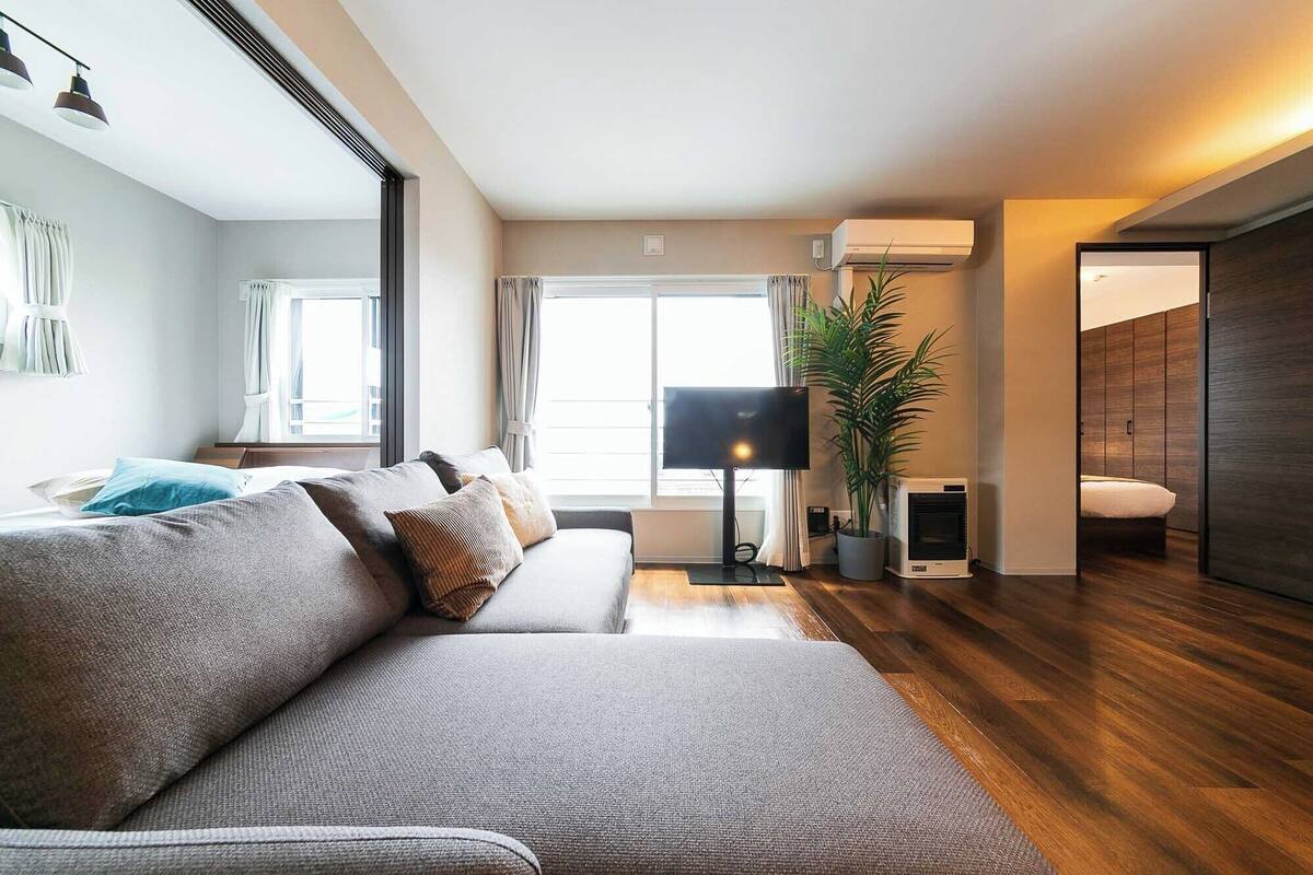A modern living room with a grey sofa, a flat-screen TV, a potted plant, and a wooden floor.