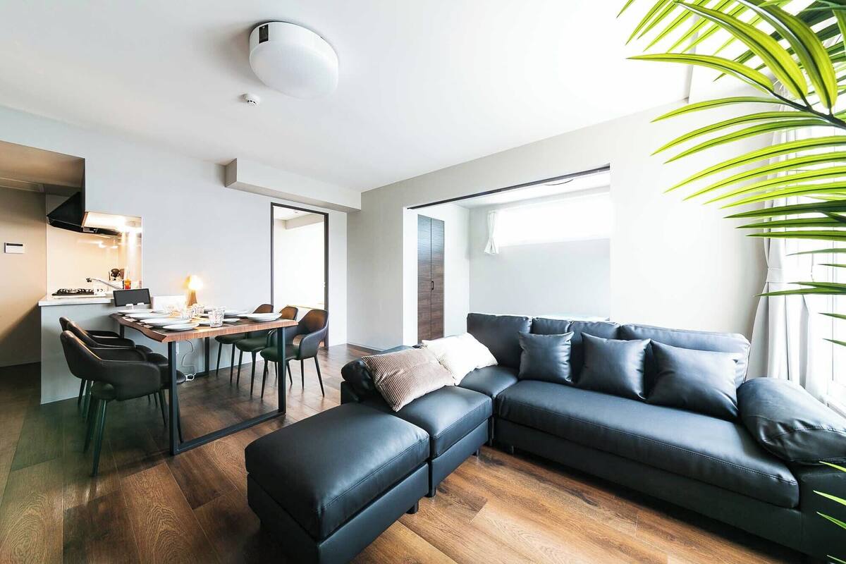 A modern living room with a black leather sofa, a dining area with a wooden table and chairs, and a kitchen with a stove and cabinets.