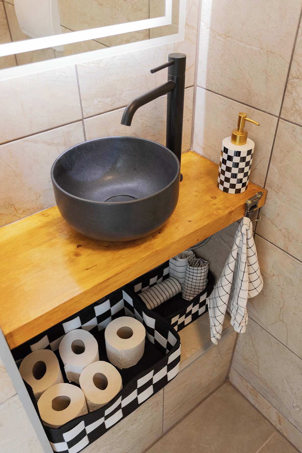 Ein Badezimmer mit einem schwarzen Waschbecken, einem karierten Seifenspender und einem Regal mit aufgerollten Handtüchern und Toilettenpapier.