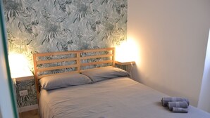 Un lit bien fait, avec une tête de lit en bois, deux lampes de chevet et un papier peint orné de motifs de feuilles tropicales.