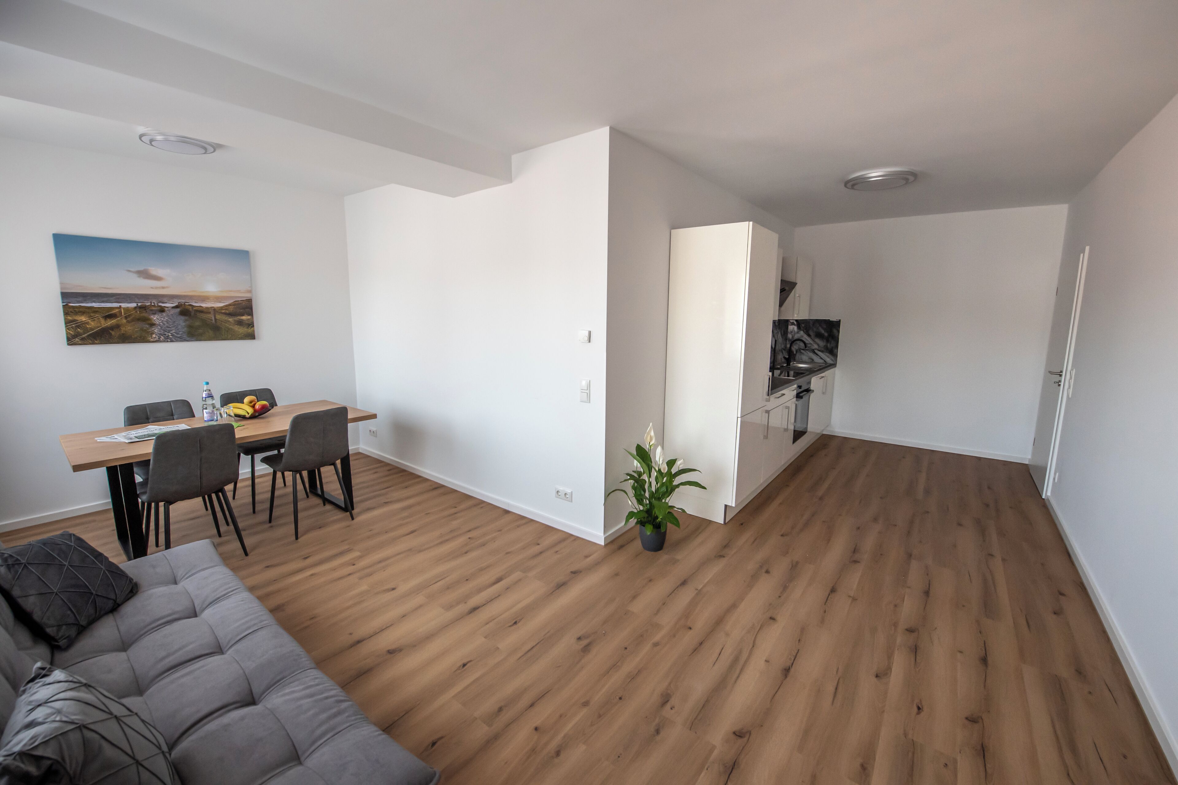 Una sala de estar moderna con piso de madera, un sofá gris, un área de comedor con mesa y sillas, y un área de cocina con armarios y un refrigerador.