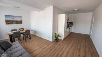 Una sala de estar moderna con piso de madera, un sofá gris, un área de comedor con mesa y sillas, y un área de cocina con armarios y un refrigerador.