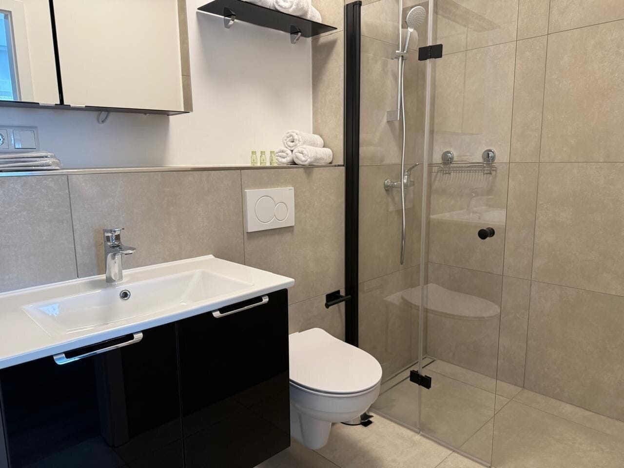 Ein modernes Badezimmer mit einem weißen Waschbecken, schwarzen Schränken, einer weißen Toilette und einer Duschkabine aus Glas.