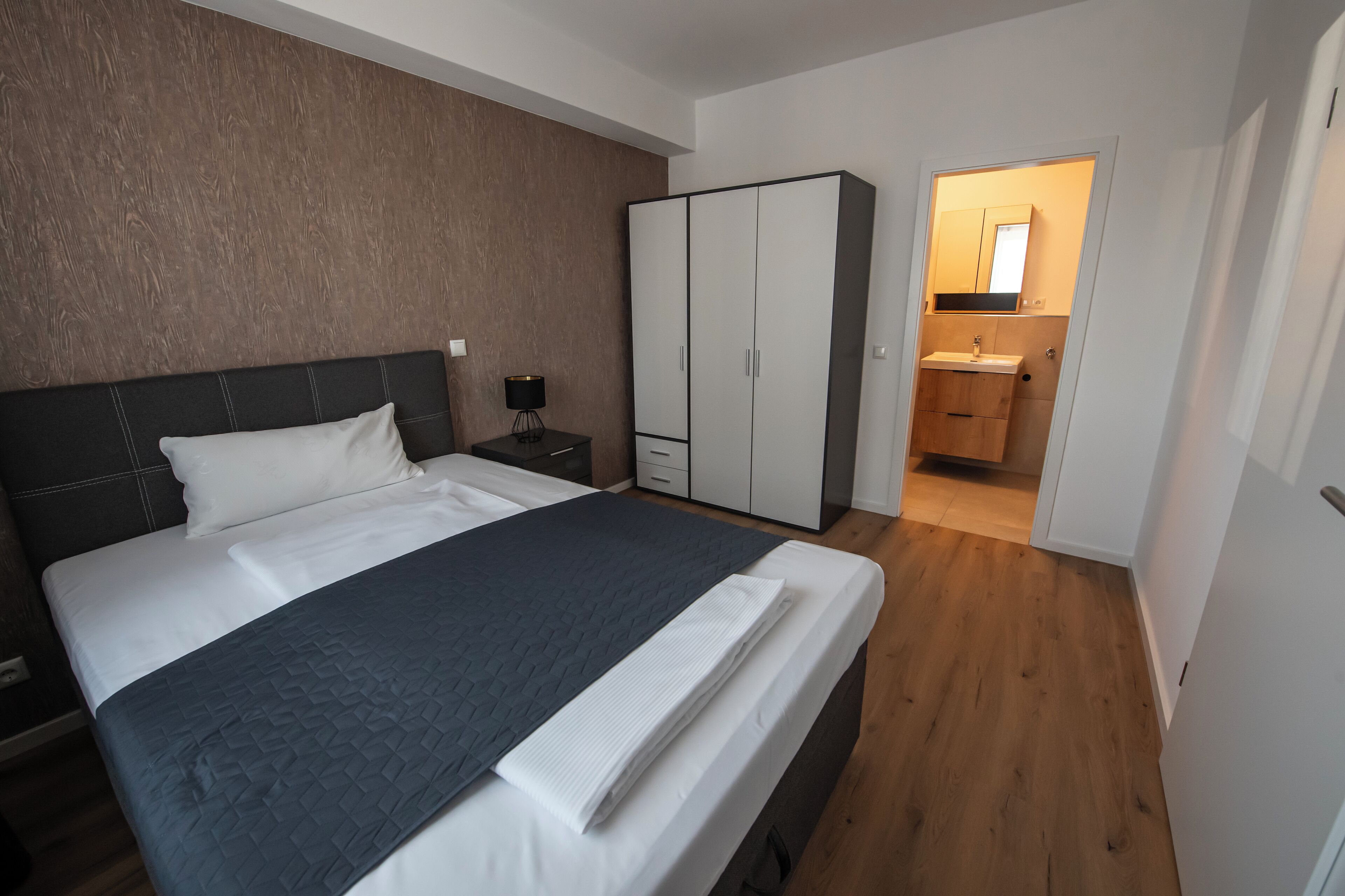 Ein Schlafzimmer mit Bett, Schrank, Nachttisch und einem durch eine offene Tür einsehbaren Badezimmer.