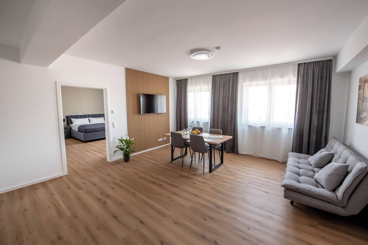 Ein modernes Wohnzimmer mit Holzboden, einer grauen Couch, einem Esstisch mit Stühlen, einem an der Wand montierten Fernseher und einem Bett im angrenzenden Zimmer.