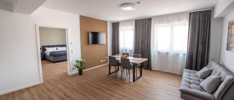 A modern living room with a wooden floor, a grey sofa, a dining table with chairs, a TV mounted on the wall, and a bed in the adjacent room.
