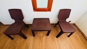 A corner room with two wicker chairs and a small table, a window with wooden frame, and wooden flooring.