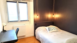 A bedroom with a bed, a desk, and a chair. There is a window with blinds, a radiator, and a wall-mounted light fixture. - In the Clouds - Free parking -Independent entrance (Mulhouse)