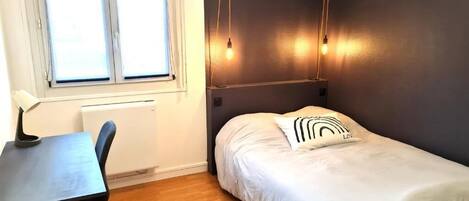 Un dormitorio con cama, escritorio y silla. Hay una ventana con persianas, un radiador y una lámpara de pared.