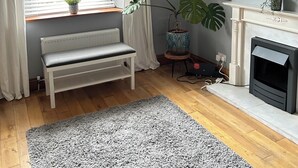 A living room with a bench, a fireplace, a large window with curtains, a potted plant, and a framed picture on the wall.