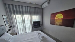 A hotel room with a large bed, a television, a painting of a sunset, and a window with curtains. - STUDIO JUST ON THE BEACH (Katerini)