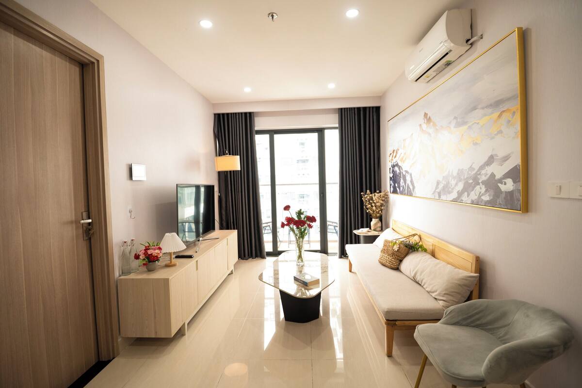 A modern hotel room with a wooden door, a flat-screen TV, a marble coffee table with a vase of flowers, a sofa with a cushion, and a large abstract painting on the wall.