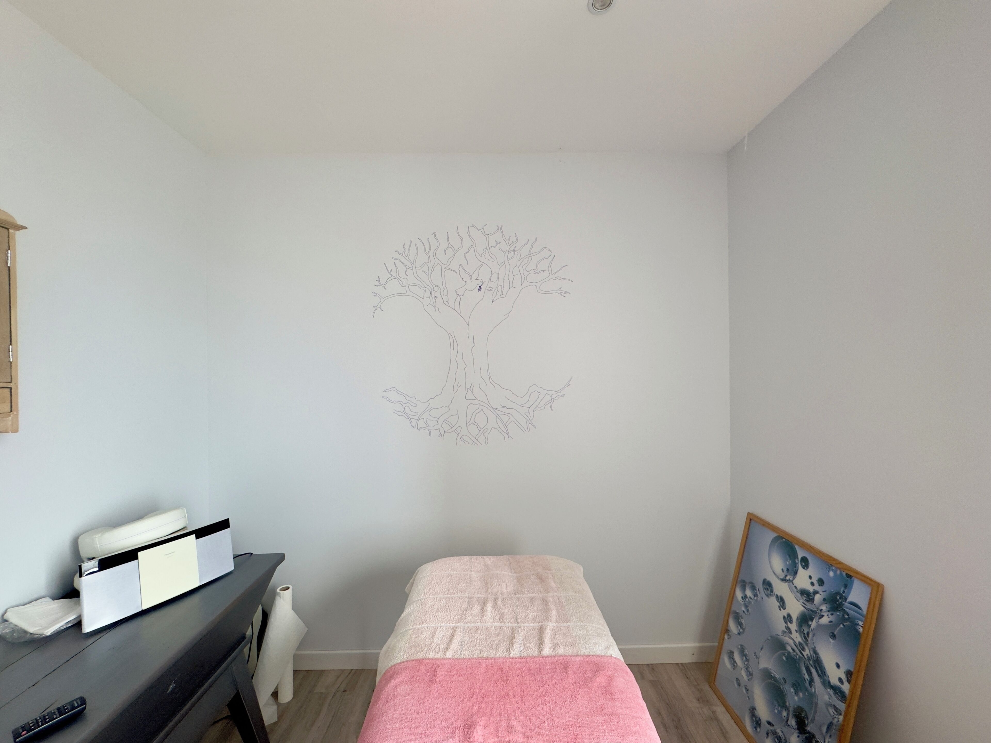 A room with a bed, a desk, a chair, a painting, and a wall with a tree drawing.