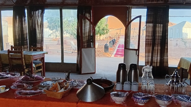 Zona de menjador amb taula preparada per a una posta, vista del paisatge desèrtic a través d'una porta oberta, i una visió de corró d'estructura amb tendes a la distància.