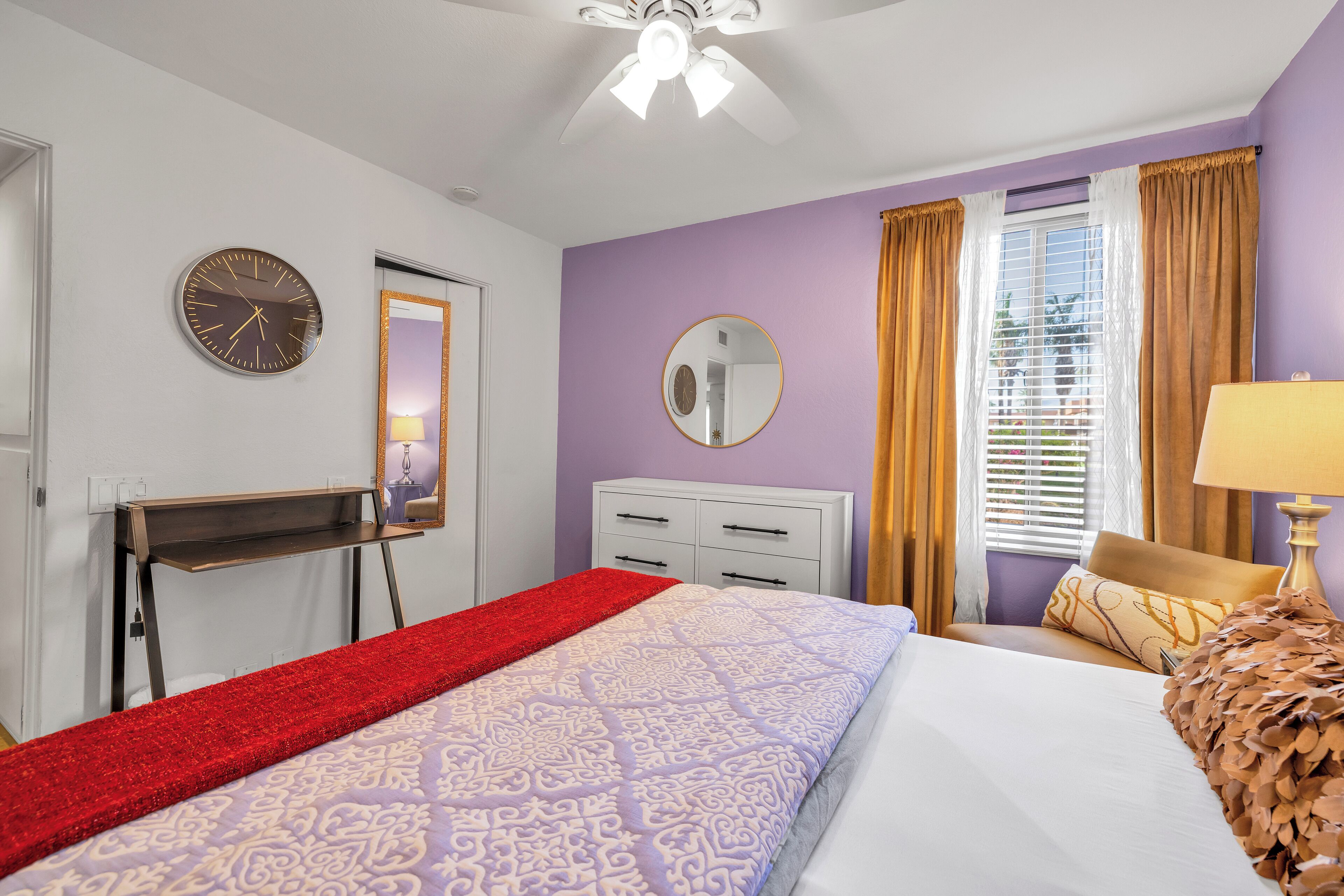 Un dormitorio con cama, escritorio, espejo, ventana con cortinas y ventilador de techo.