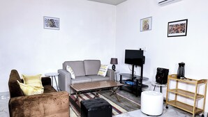 A living room with a grey sofa, a brown armchair, a coffee table, a TV on a stand, a lamp, a speaker, and a small shelf.