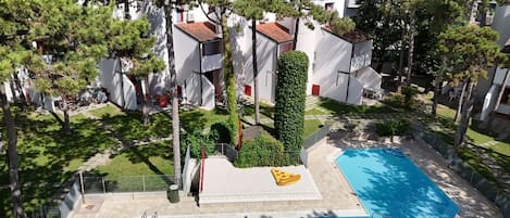 Una piscina en la azotea decorada con una porción de pizza, rodeada de árboles y edificios blancos con detalles rojos.