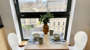 A dining area with a table set for two, a vase with a plant, and a view of buildings through a window.