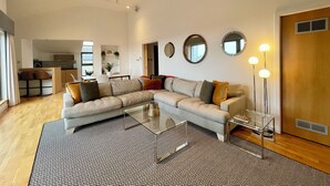 A modern living room with a sectional sofa, a glass coffee table, and a herringbone-patterned rug. There are three wall-mounted mirrors and a floor lamp.