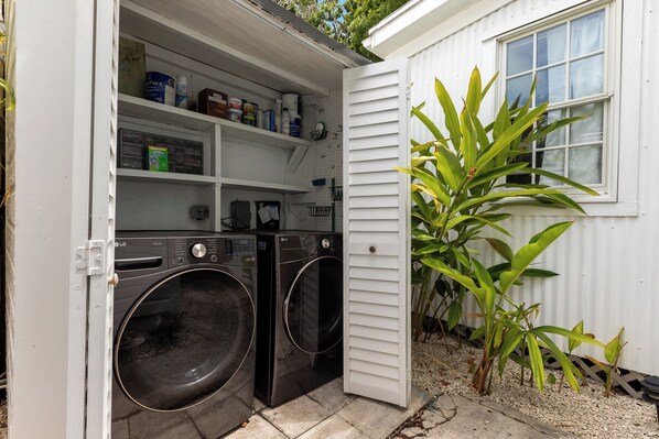 A laundry room with two front-loading washers and dryers, a shelf with cleaning supplies, and a small plant outside.