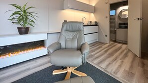 Ein modernes Wohnzimmer mit einem grauen Ledersessel, einem kleinen Topfpflanzen, einem eingebauten Kamin und einer Küchenzeile mit weißen Schränken und einer Arbeitsplatte.