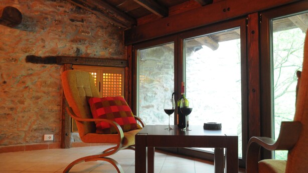 A room with a stone wall, a wooden ceiling, and a large window. There is a chair with a red cushion, a table with a bottle and glasses, and a small rug on the floor.