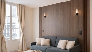 A modern living room with a grey sofa, a wooden accent wall, a ceiling fan, and a window with curtains. - 1608 Saint-Honoré (Paris)