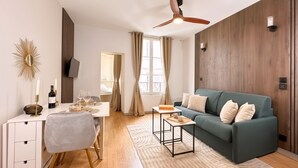 A modern living room with a sofa, a dining table, a TV mounted on the wall, and a ceiling fan. - 1608 Saint-Honoré (Paris)