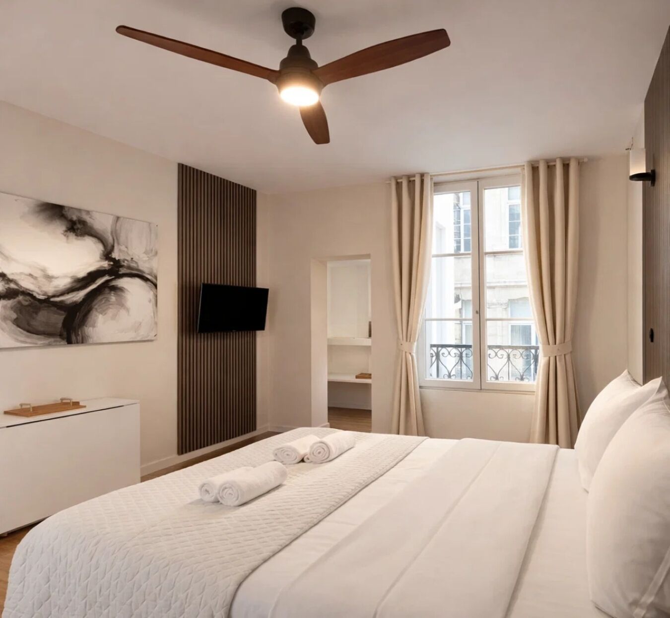 Ein Schlafzimmer mit Bett, Deckenventilator, an der Wand montiertem Fernseher, Fenster mit Vorhängen und einem Bild an der Wand.