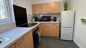 A compact kitchen with wooden cabinets, a black stove, a microwave, a coffee maker, and a refrigerator. - Two Bedroom Cottage in Lumsden - Contractors (Huntly)